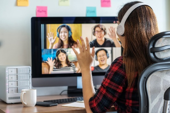 A young with her back to the camera, sitting in a room at a desk wearing headphones, looking at her open laptop in front of her